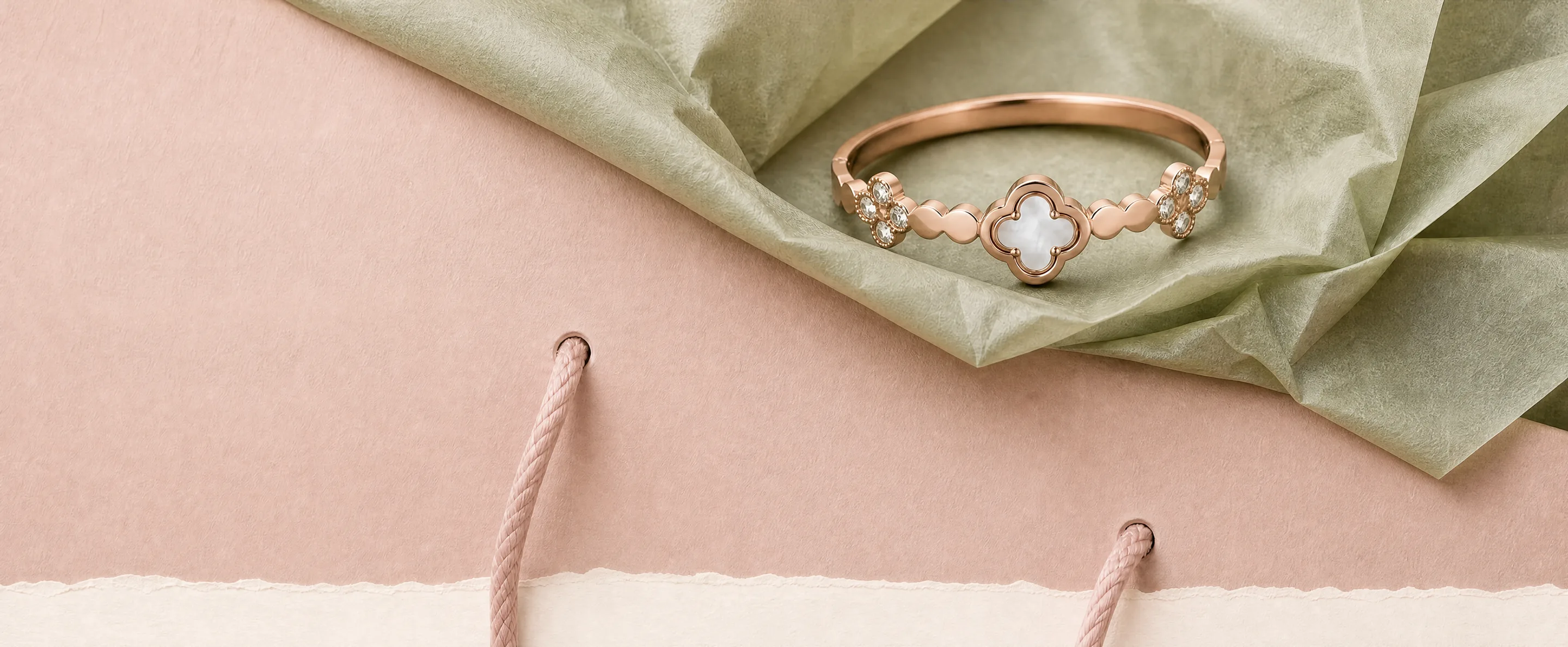 A rose-gold clover bangle with a mother-of-pearl centre resting in sage tissue paper on a blush gift bag